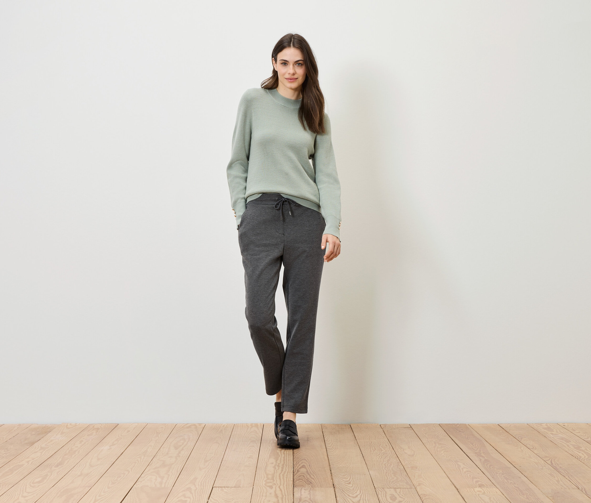 Eine Frau steht auf einem Holzboden vor einer weißen Wand. Sie trägt einen grünen Pullover und graue Hosen.