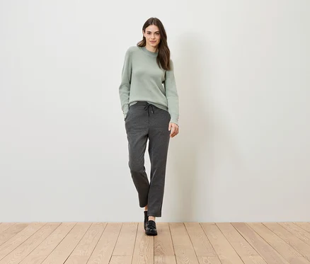 Eine Frau steht auf einem Holzboden vor einer weißen Wand. Sie trägt einen grünen Pullover und graue Hosen.