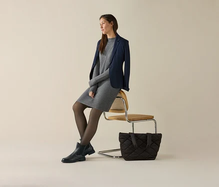 Frau sitzt auf einem Stuhl, trägt ein graues Kleid und einen marineblauen Jerseyblazer. Neben dem Stuhl steht eine Handtasche.