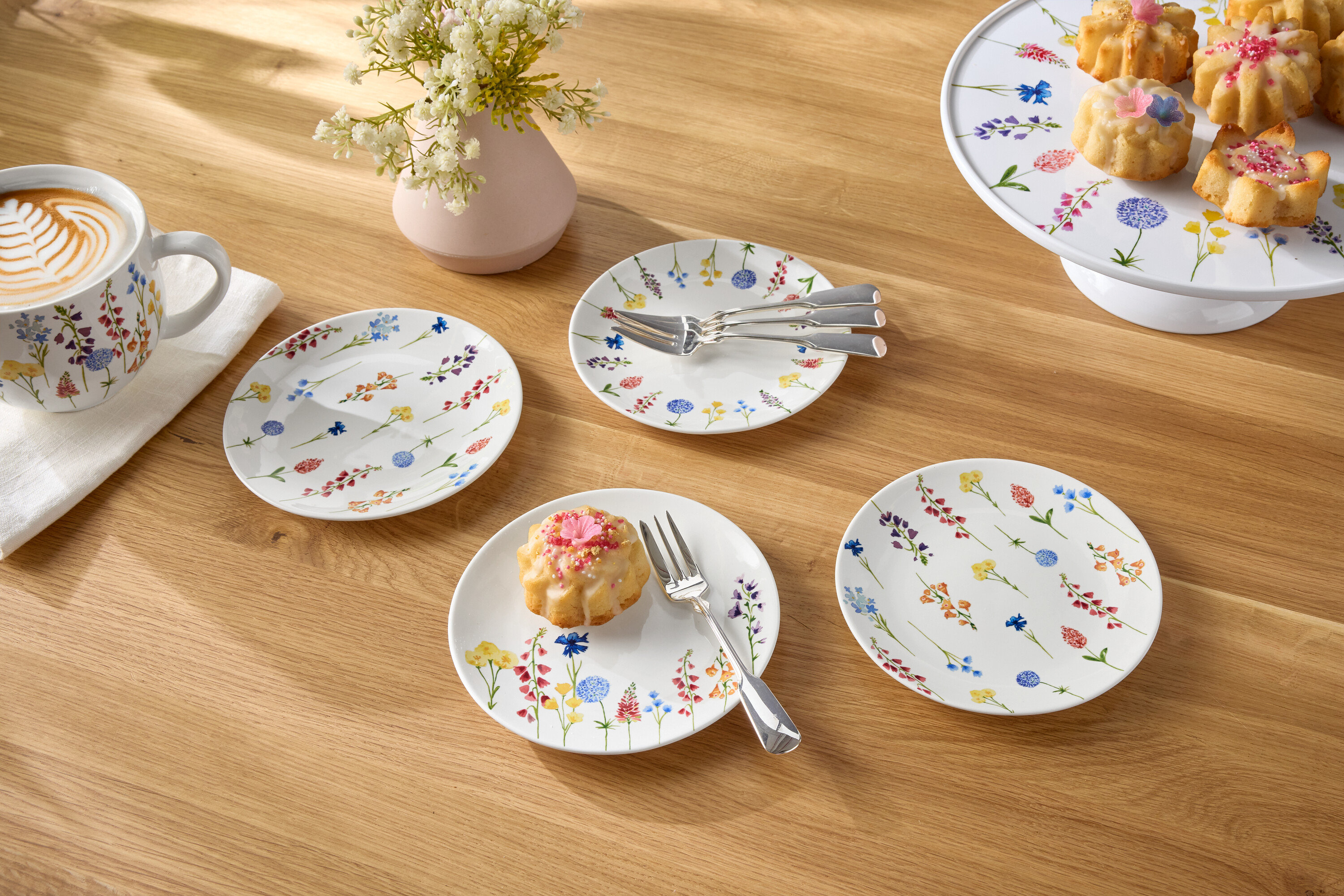 Auf einem Holztisch stehen eine Tasse Kaffee, eine Blumenvase, Teller und kleine Kuchen.