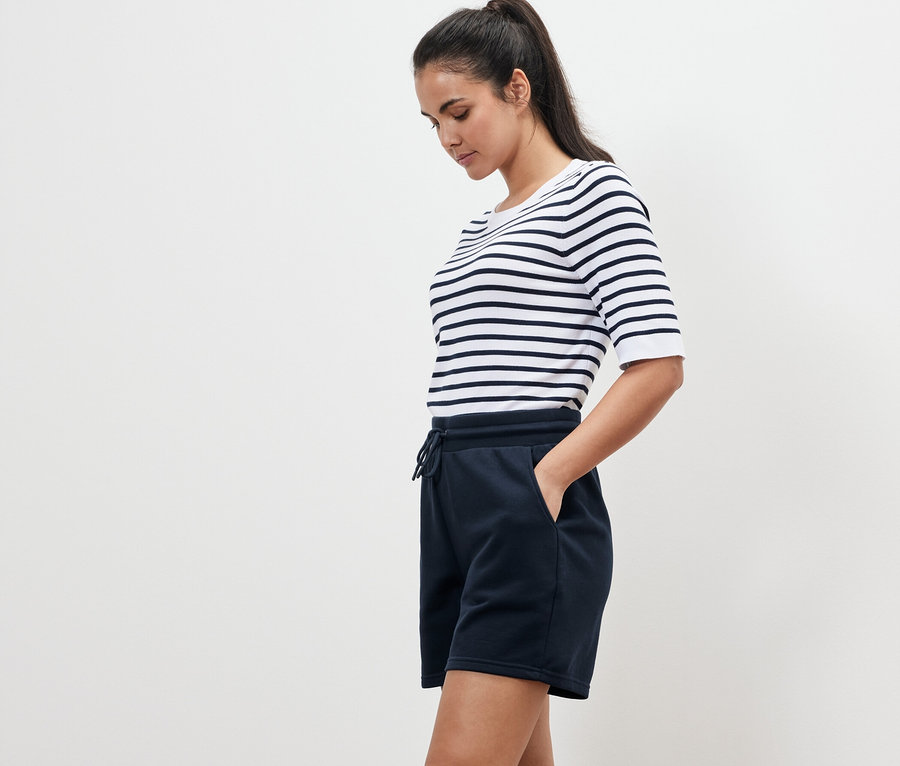 Frau mit gestreiftem T-Shirt und marineblauen Sweatshorts steht seitlich.