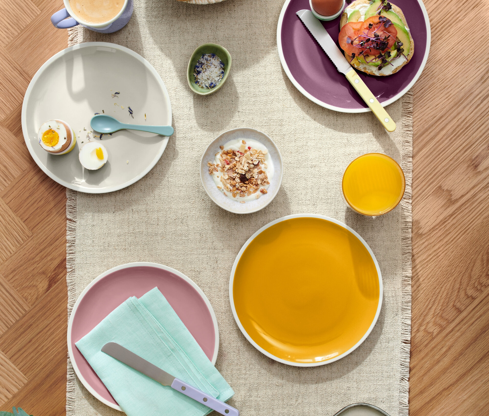 Draufsicht auf einen gedeckten Tisch mit verschiedenen Speisen, darunter Gebäck in einem Korb, Teller mit Eiern, Avocado-Toast und Käse, zusammen mit Getränken wie Kaffee und Saft.