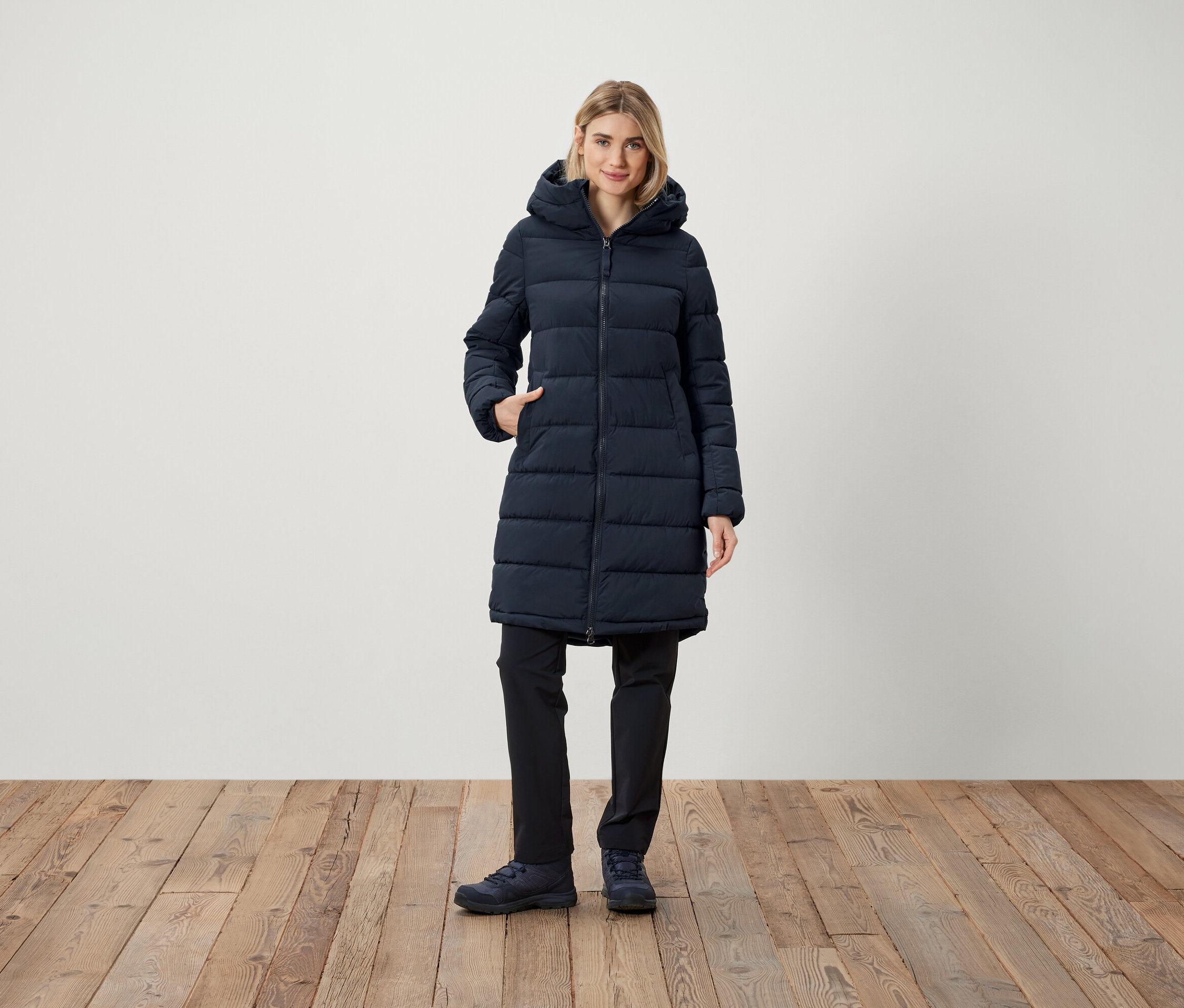 Frau steht mit der Hand in der Tasche einer dunkelblauen Steppjacke mit Kapuze.