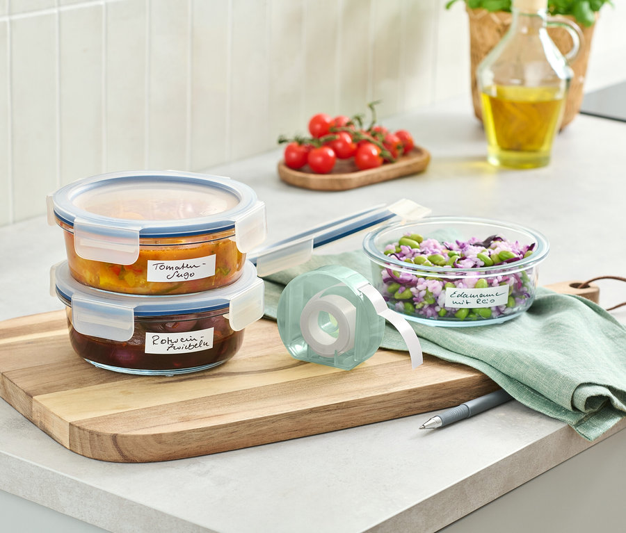 Zwei Glasbehälter mit Essen gestapelt auf einem Holzbrett neben einer Etikettenrolle.