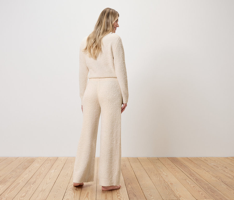 Frau steht mit dem Rücken zur Kamera, gekleidet in einen Pullover und eine weite Hose aus Bouclé-Strick.