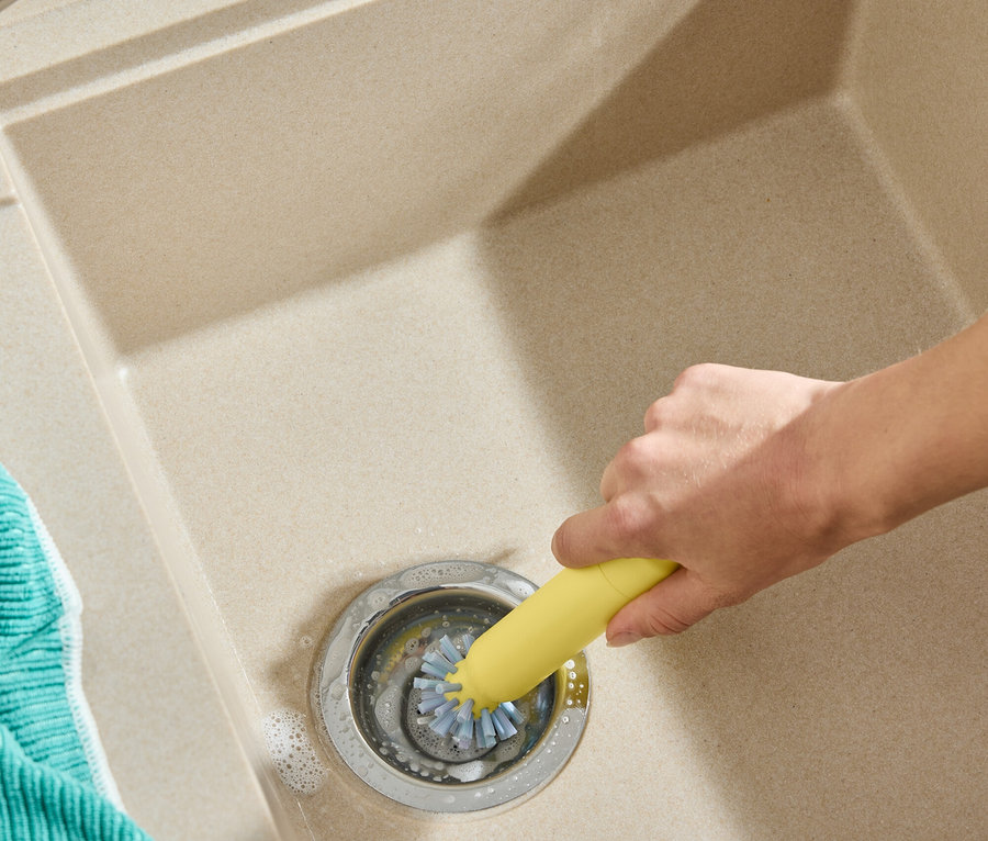 Eine Hand reinigt einen Spülenabfluss mit einer gelben Bürste. Links liegt ein blaugrünes Tuch.