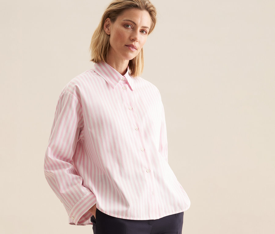Blonde Frau trägt eine roséfarbene, gestreifte Seidensticker Oversize Hemdbluse und dunkle Hose, Hände in den Taschen.