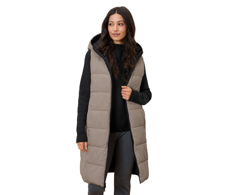 Frau steht und trägt eine lange, gesteppte Weste mit Kapuze.