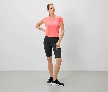 Frau steht in einem rosa T-Shirt, schwarzen Radlerhosen und Turnschuhen da und posiert.