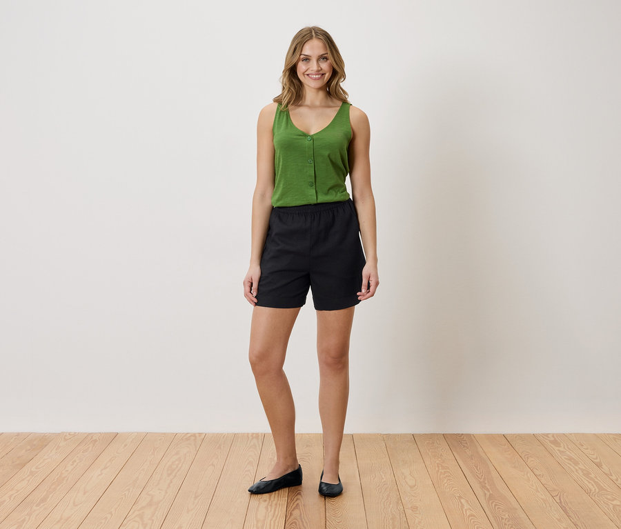 Frau mit grünem Top und schwarzen Shorts steht auf einem Holzboden vor einer weißen Wand.