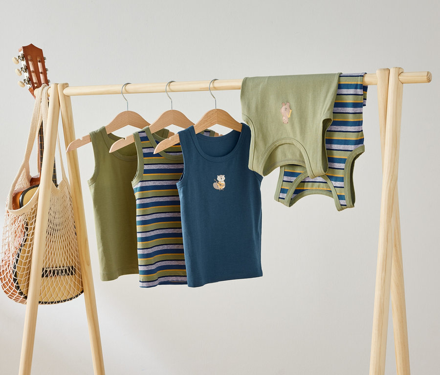 Auf einer Holzstange hängen vier Kinder Tanktops in Grün, gestreift und Blau mit Tiermotiven. Eine Gitarre hängt in einer Netztasche an der Stange.