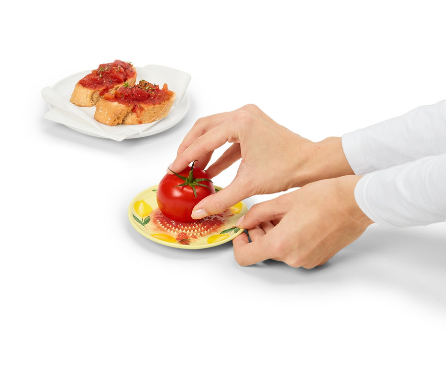 Hände halten einen gelben Teller mit einer roten Tomate. Im Hintergrund befinden sich zwei Tomatenbrote auf einem weißen Teller.