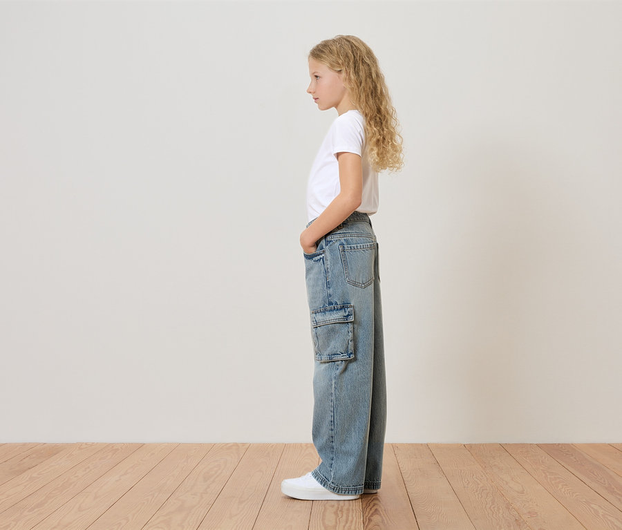 Ein junges Mädchen mit blonden, gelockten Haaren steht seitlich und trägt ein weißes T-Shirt, weite Jeans mit Seitentasche und weiße Turnschuhe.