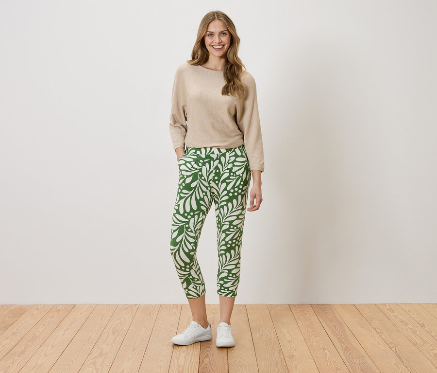 Frau steht mit den Händen in den Taschen vor einer Wand. Sie trägt einen beigefarbenen Pullover und eine grüne Hose mit weißem Blattmuster.