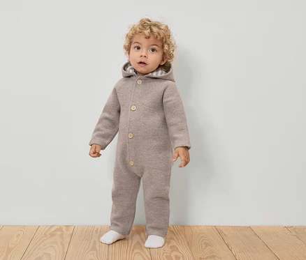 Ein Kleinkind mit lockigen Haaren trägt einen hellbraunen Woll-Overall mit Kapuze und Knöpfen, dazu weiße Socken.