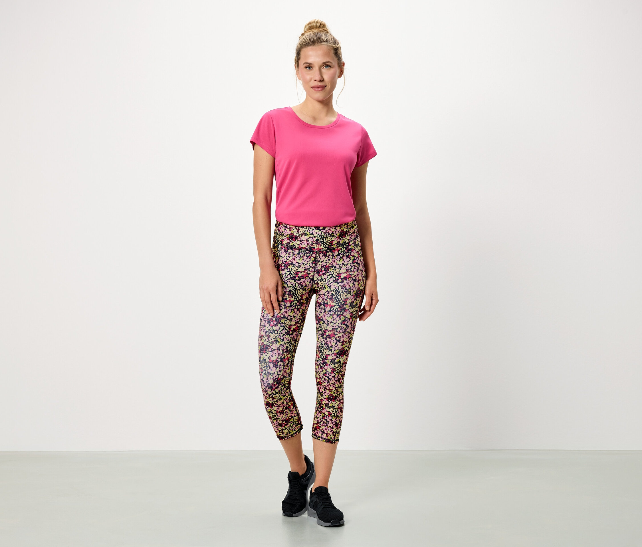 Frau steht vor einem weißen Hintergrund und trägt ein pinkes T-Shirt und blumige Leggings.