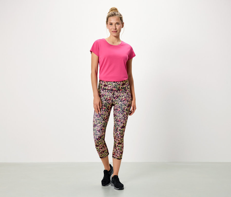 Frau steht vor einem weißen Hintergrund und trägt ein pinkes T-Shirt und blumige Leggings.