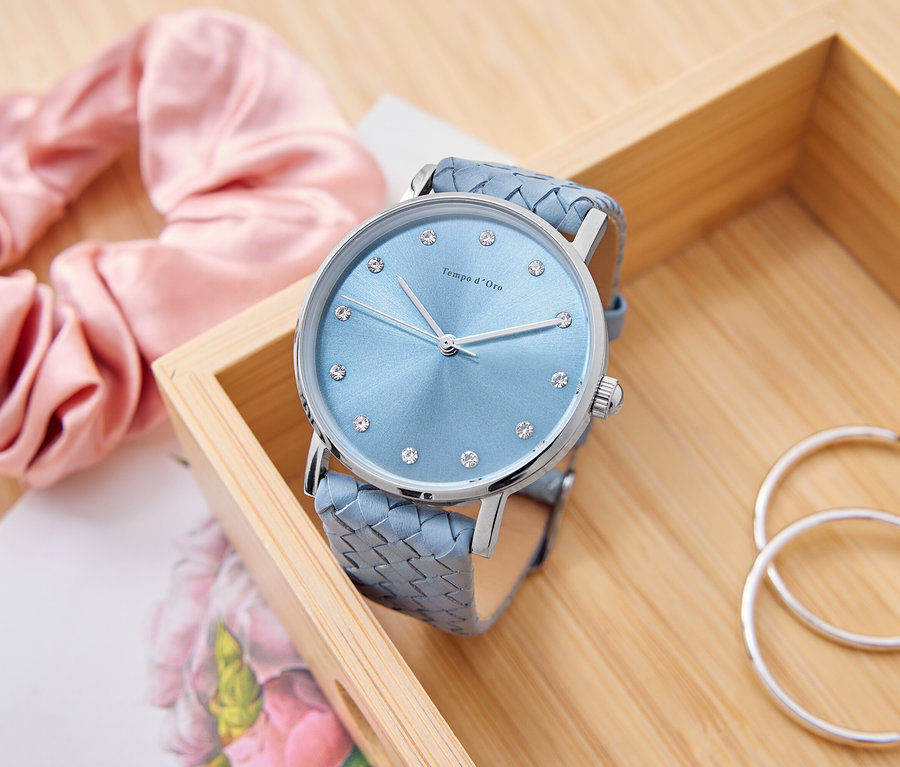 Eine hellblaue Uhr mit Strasssteinen liegt in einer Holzkiste, daneben ein rosa Scrunchie, ein Notizblock und silberne Creolen.