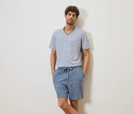 Ein Mann mit lockigem Haar, der ein gestreiftes T-Shirt und blaue Shorts trägt, steht vor einer weißen Wand.