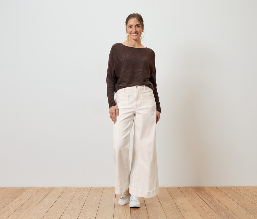 Eine Frau steht vor einer Wand und trägt einen braunen Pullover, eine weite, helle Hose und weiße Schuhe.