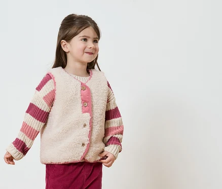 Lächelndes Mädchen mit gestreiftem Pullover und beiger Weste mit rosa Akzenten vor einem weißen Hintergrund.