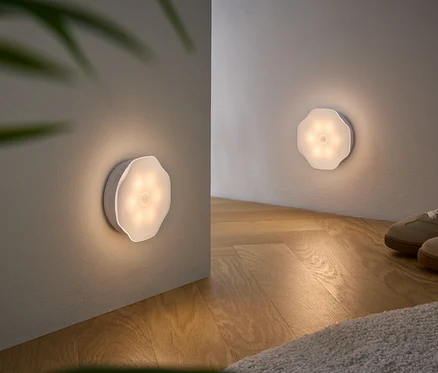 Zwei an Wänden montierte, blütenförmige, weiße Lichter beleuchten einen Raum mit Holzboden und beigen Schuhen.