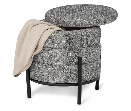 Ein grauer, runder Hocker mit schwarzem Beingestell und einer beigen Decke darauf.