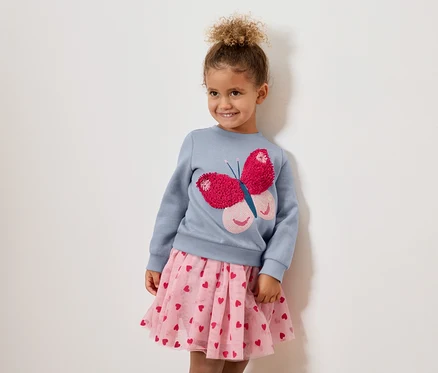 Mädchen mit lockigen Haaren trägt ein blaues Sweatshirt mit einem pinkfarbenen Schmetterling und einen rosa Tüllrock mit Herzen.