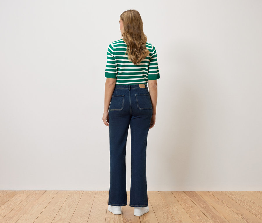 Rückansicht einer Frau mit langem, gewelltem Haar, die ein grün-weiß gestreiftes Oberteil und dunkelblaue Jeans mit ausgestelltem Bein und weißen Turnschuhen trägt.