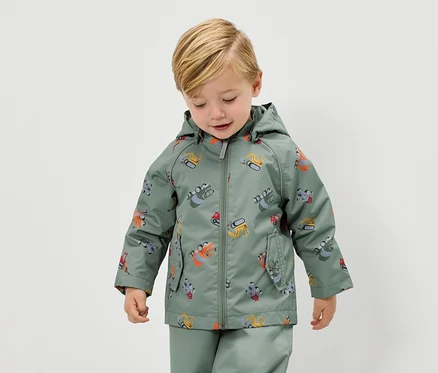 Ein blonder Junge trägt eine grüne Regenjacke mit Bagger-Motiv und eine passende Regenhose.
