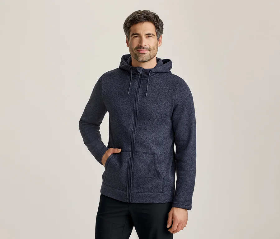 Ein Mann in einer dunkelblauen Kapuzenjacke mit Reißverschluss, eine Hand in der Tasche, vor hellem Hintergrund stehend.