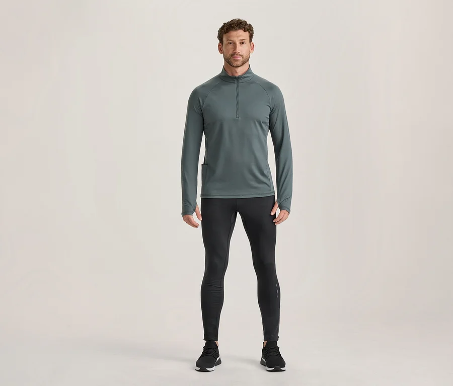 Mann steht in einem dunkelgrauen Langarm-Sportoberteil mit Reißverschluss und Daumenlöchern sowie schwarzen Sportleggings und schwarzen Sportschuhen.