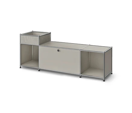 Niedriges, modulares Möbelstück mit Metallrahmen und hellgrauen Paneelen. Links ein perforierter Korb über einem offenen Fach, mittig ein geschlossener Schrank, rechts ein offenes Fach.