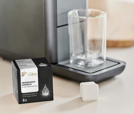 Eine Packung Qbo Reinigungskapseln steht neben einer Kaffeemaschine und einem Glas.
