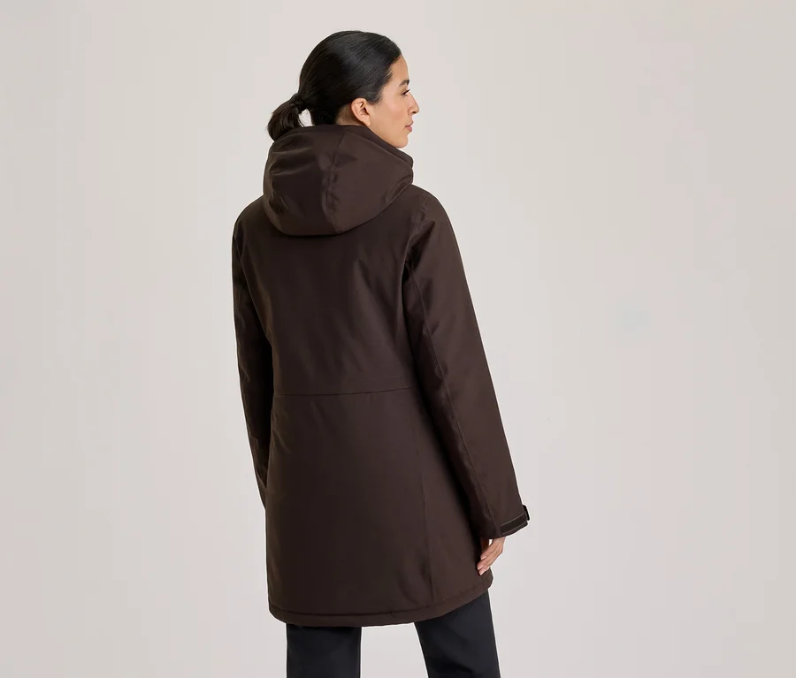 Frau von hinten, leicht nach rechts gedreht, trägt einen dunkelbraunen Kapuzenparka und dunkle Hose. Ihr Haar ist zu einem Pferdeschwanz gebunden.