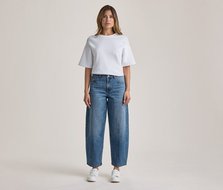 Stehende Frau in einem weißen T-Shirt, blauen weiten Jeans und weißen Sneakern.