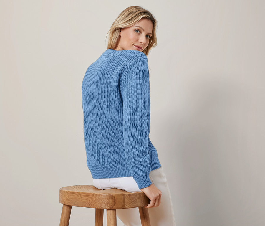 Eine Frau mit blonden Haaren trägt einen blauen Grobstrick-Cardigan und eine weiße Hose, sitzt auf einem Holzschemel und blickt über ihre linke Schulter.