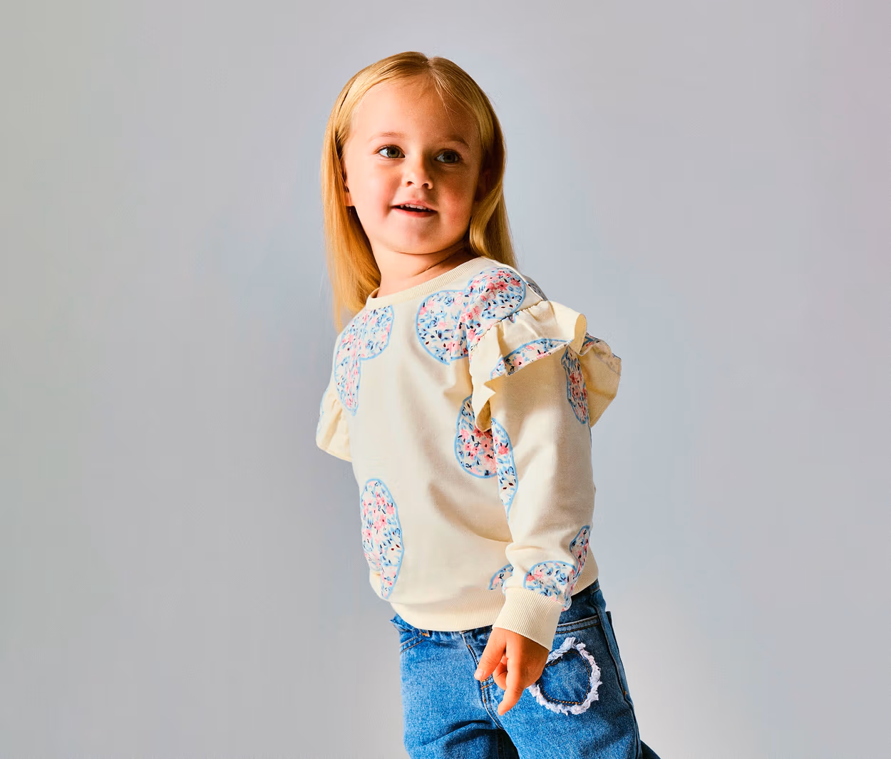 Junges Mädchen mit blondem Haar trägt einen cremefarbenen NAME IT Kinder-Sweater mit floralen Herzmustern und Rüschen an den Schultern, dazu blaue Jeans, blickt nach rechts.
