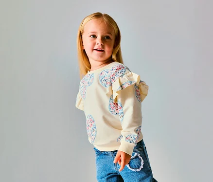 Junges Mädchen mit blondem Haar trägt einen cremefarbenen NAME IT Kinder-Sweater mit floralen Herzmustern und Rüschen an den Schultern, dazu blaue Jeans, blickt nach rechts.