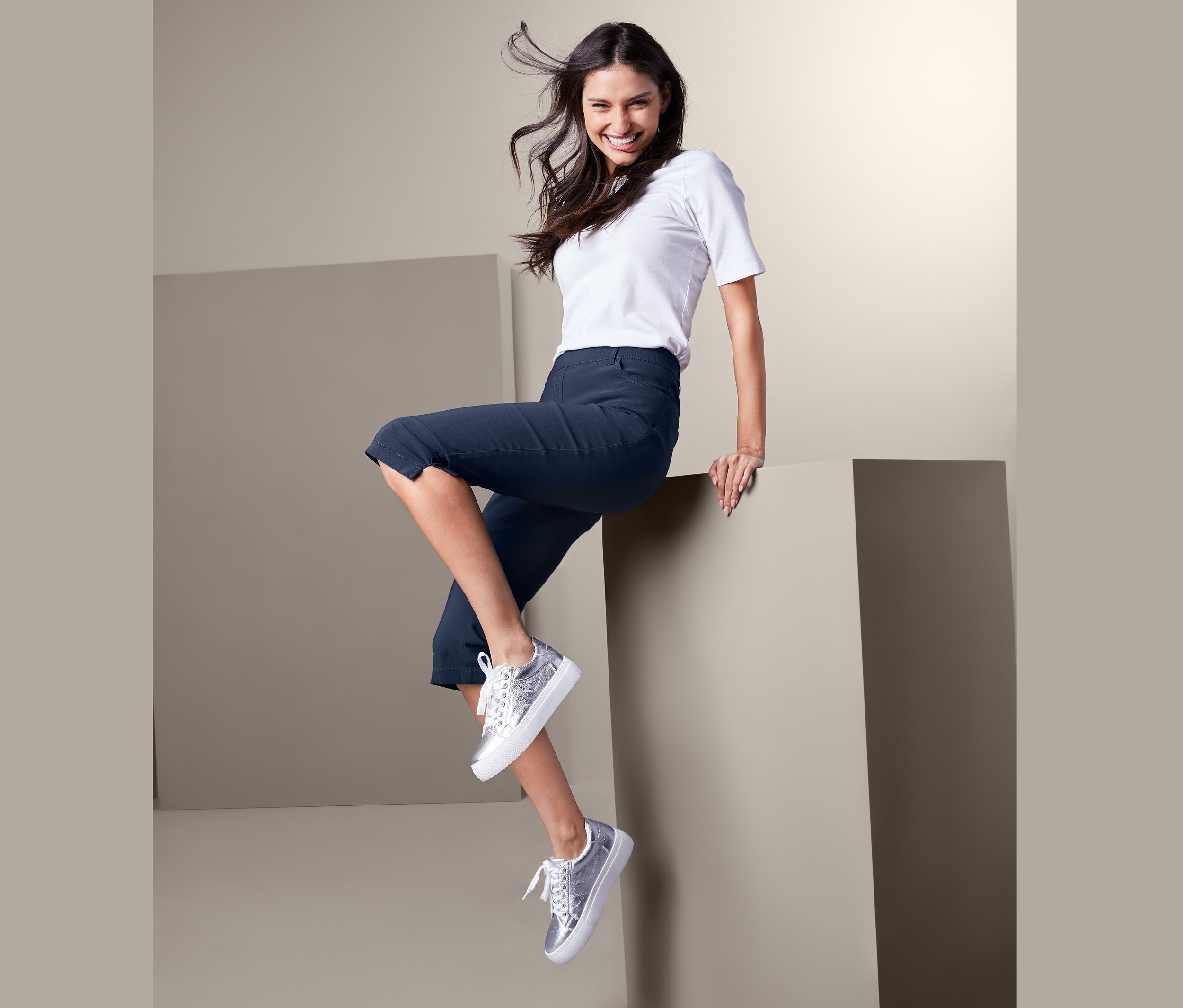 Eine lächelnde Frau sitzt auf der Kante eines Würfels und trägt ein weißes T-Shirt und eine dunkelblaue Bengalin-Hose in 3/4-Länge.