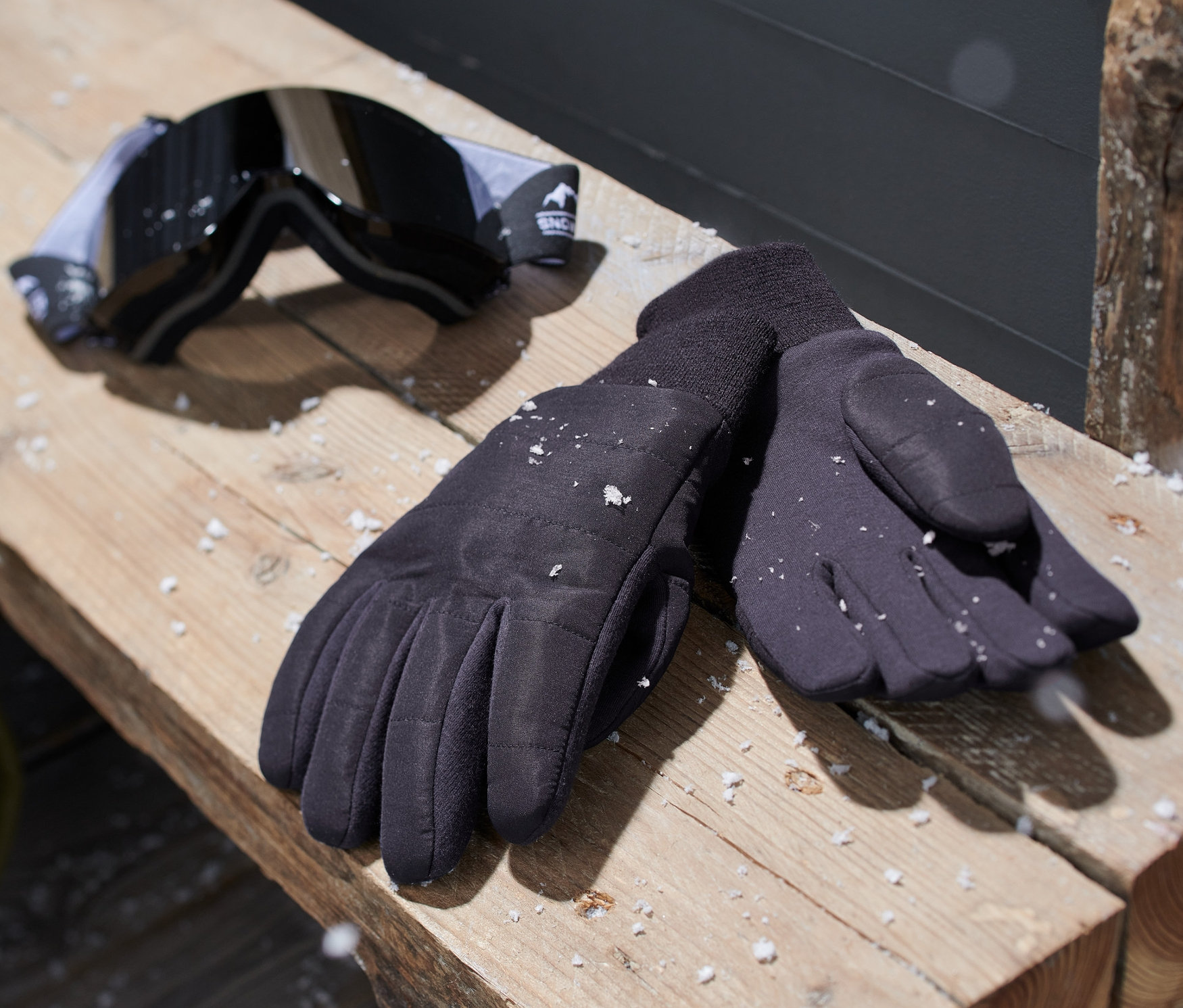 Schwarze Strickhandschuhe und eine Skibrille liegen auf einer Holzbank.