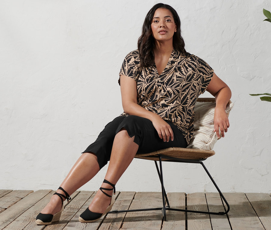 Frau sitzt auf einem Stuhl und trägt Espadrilles mit Keilabsatz, eine 3/4-Stretchhose und ein bedrucktes Shirt mit Stehkragen.