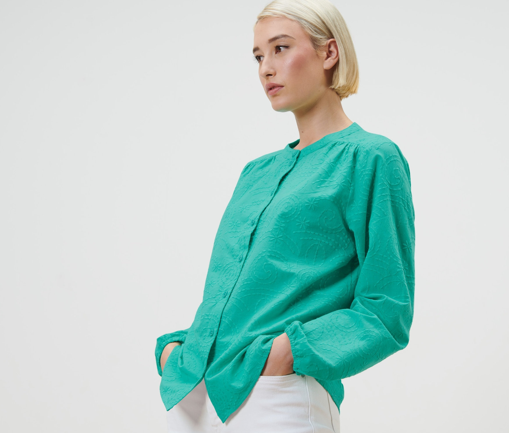 Blonde Frau posiert in grüner Bluse mit Stickerei und weissen Hosen.