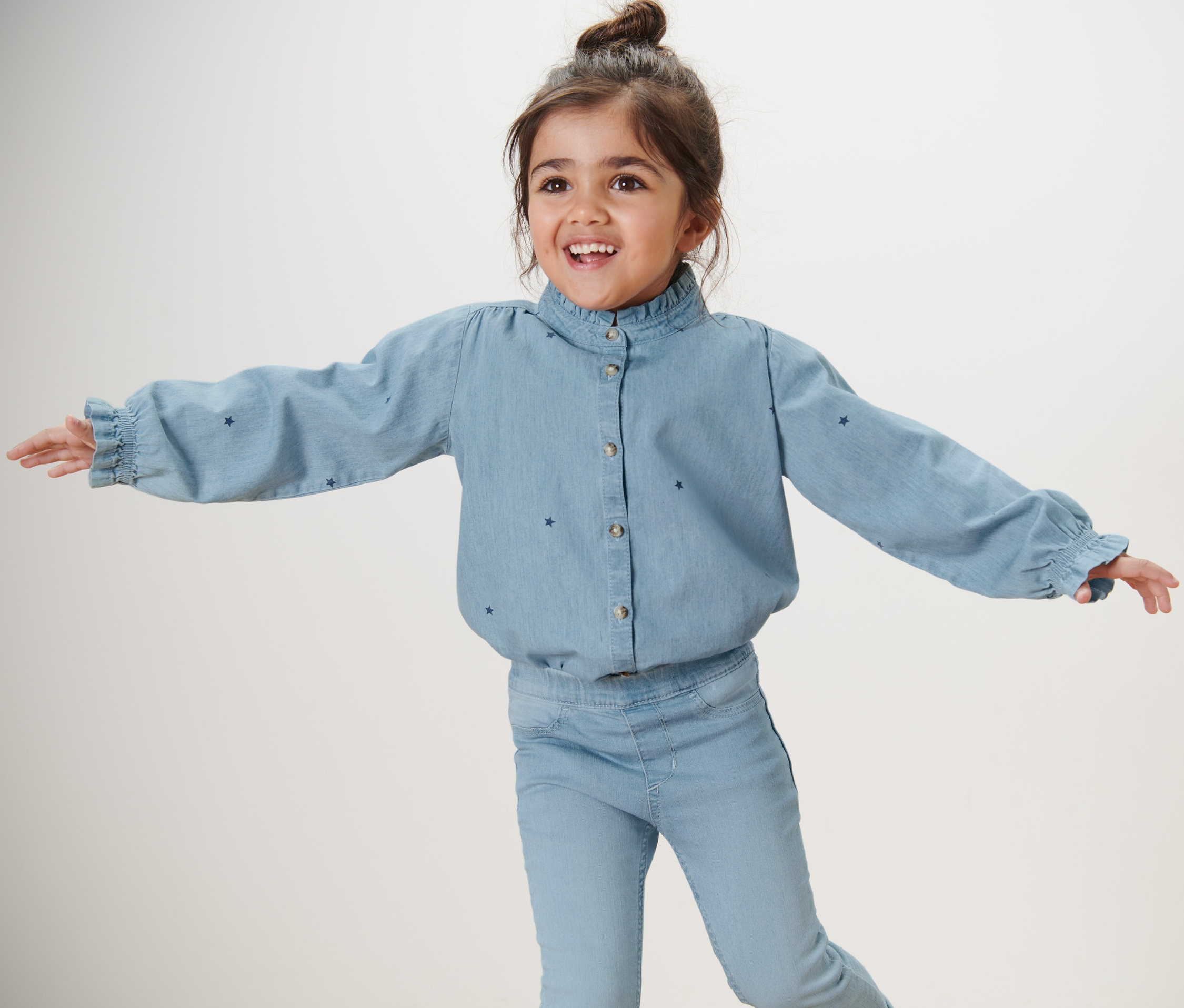 Lächelndes Mädchen mit ausgestreckten Armen trägt eine Kinder-Chambray-Bluse und Jeans.