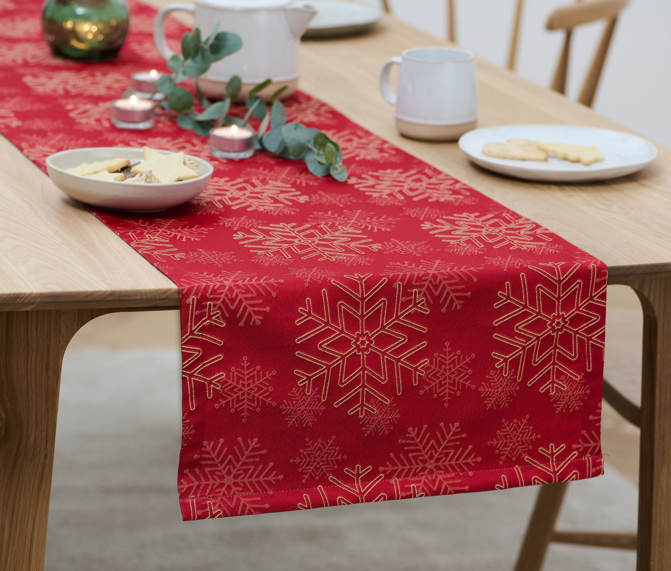 Auf einem Holztisch liegt ein roter Jacquard-Tischläufer mit Schneeflockenmuster, auf dem Teller mit Keksen, Kerzen und eine Teekanne stehen.