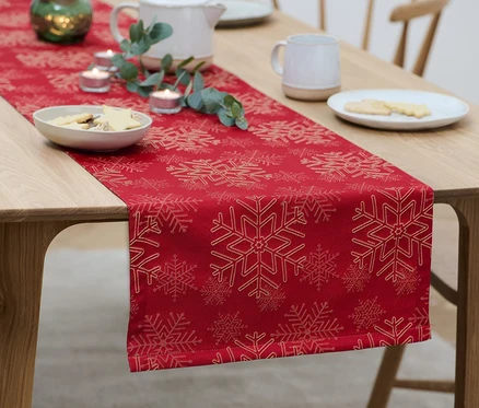 Auf einem Holztisch liegt ein roter Jacquard-Tischläufer mit Schneeflockenmuster, auf dem Teller mit Keksen, Kerzen und eine Teekanne stehen.