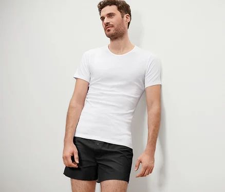 Mann lehnt an einer Wand, trägt 2 Qualitäts-Feinripp-Unterhemden und schwarze Boxershorts.