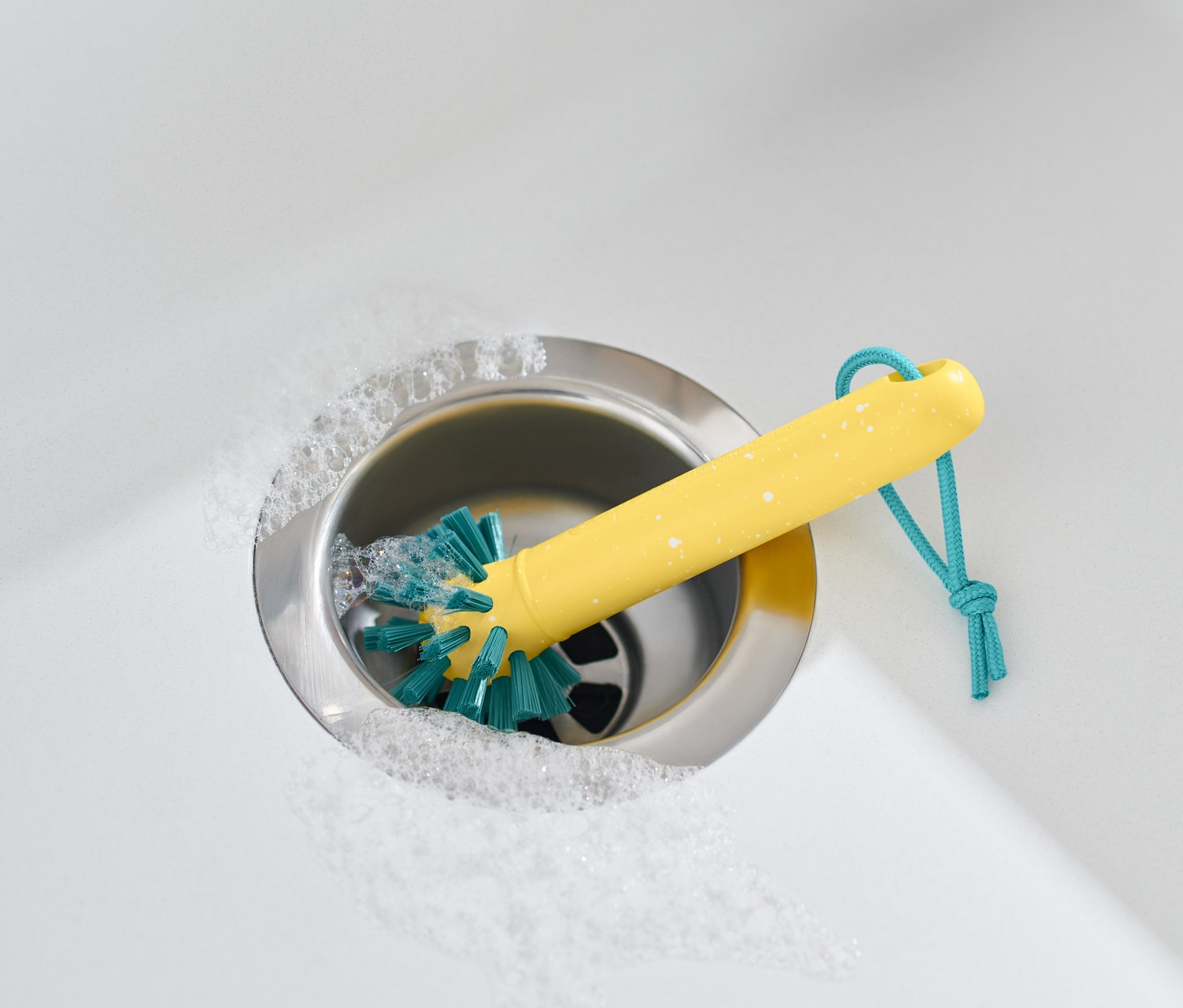 Gelbe Universalreinigungsbürste in Edelstahlspüle mit Schaum und beigem Handtuch.