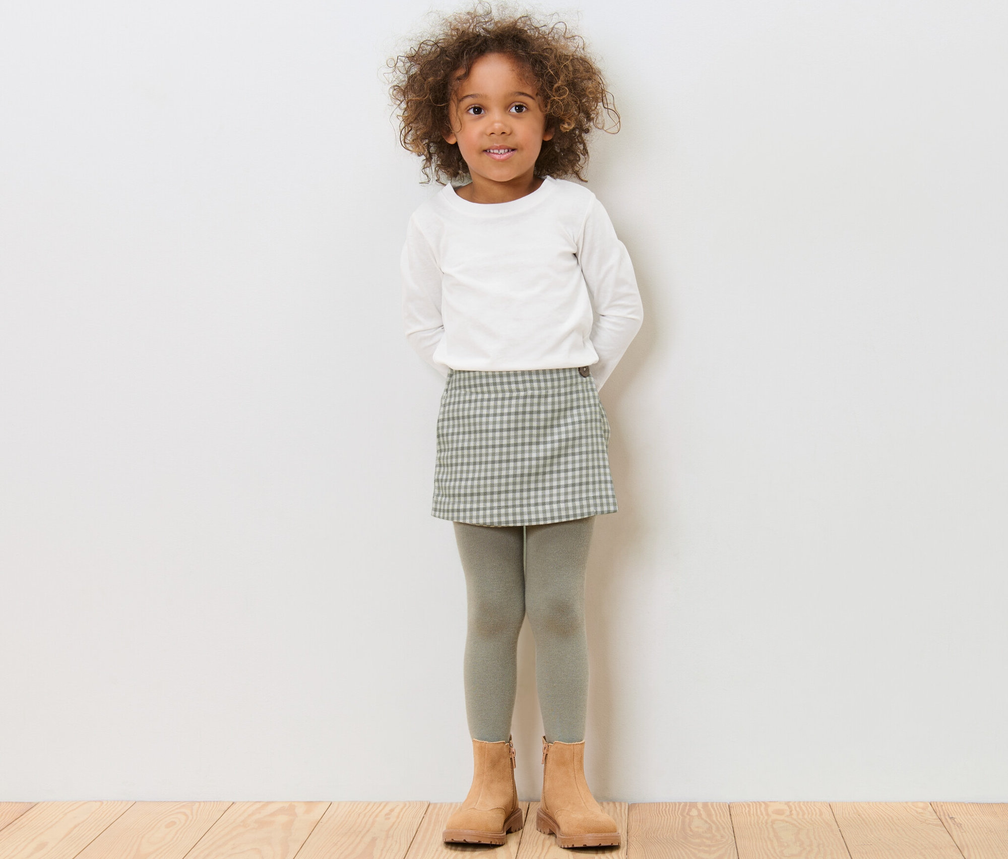 Mädchen steht und trägt einen Kinder-Hosenrock, ein weißes Oberteil und braune Schuhe.