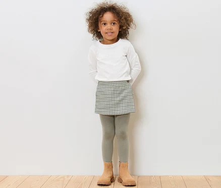 Mädchen steht und trägt einen Kinder-Hosenrock, ein weißes Oberteil und braune Schuhe.